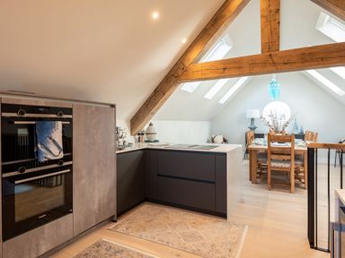 A kitchen with built in ovens and an island opposite a dining table with chairs under wooden beams at Church Rooms in Port Isaac