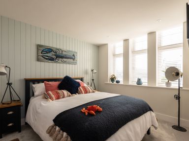 A bedroom with a bed featuring multiple pillows a blue throw blanket and a stuffed crab toy a nightstand with a lamp a large window with blinds and decorative items a lobster painting above the bed at Church Rooms in Port Isaac