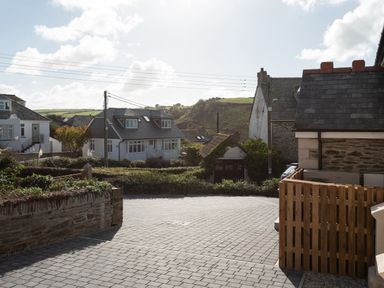 A paved driveway with wooden fencing and stone walls in a residential area with houses and green hills in the background at Church Rooms in Port Isaac