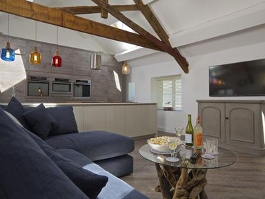 A living area with a blue sofa a glass coffee table with drinks and popcorn a TV on a gray cabinet and kitchen appliances at The Barn 20 At The Beach Torcross