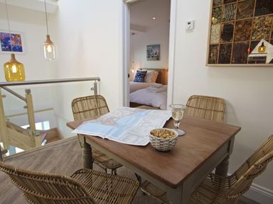 A dining area with a wooden table and four wicker chairs a map and bowl on the table a bedroom visible through doorway at The Barn 20 At The Beach Torcross