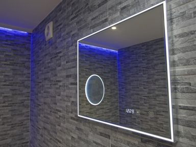 A bathroom wall with gray stone tiles and a rectangular LED mirror showing a clock at The Barn 20 At The Beach in Torcross