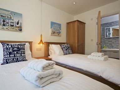 A bedroom with two single beds a wooden wardrobe and an open door showing a bathroom at The Barn 20 At The Beach in Torcross
