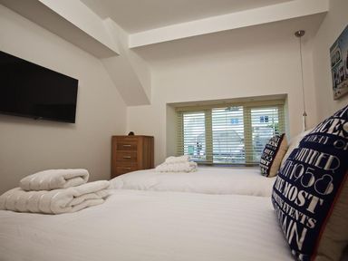 A bedroom with two beds white bedding and towels a wall mounted television and a window with blinds at The Barn 20 At The Beach in Torcross