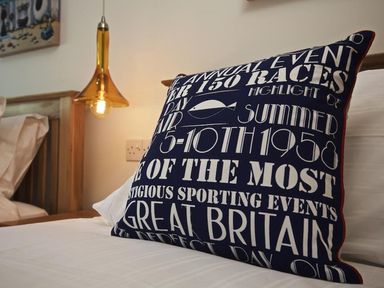 A close up of a navy blue pillow with white text on a bed with white sheets and a wooden headboard at The Barn 20 At The Beach Torcross