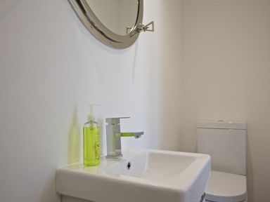 A bathroom sink with a soap dispenser and a round mirror above it a toilet in the background at The Barn 20 At The Beach Torcross