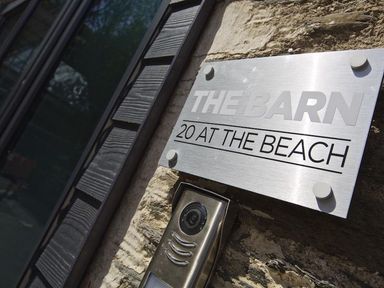 A metal sign saying THE BARN 20 AT THE BEACH mounted on a stone wall with a video doorbell below it at The Barn 20 At The Beach Torcross