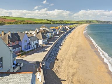 A row of houses along a concrete promenade next to a sandy beach and a body of water at The Barn 20 At The Beach in Torcross