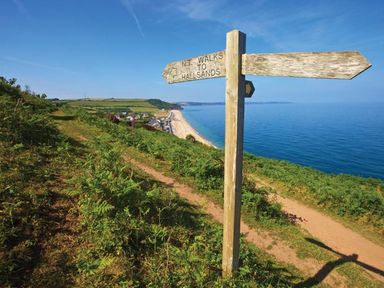 A wooden signpost on a coastal walking path overlooking green hills and a beach at The Barn 20 At The Beach in Torcross