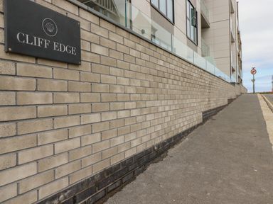 A brick wall with a sign that says Cliff Edge next to a sidewalk and a modern building at 13 Cliff Edge in Newquay