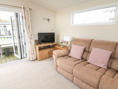 A living room with a brown sofa with pink cushions a wooden TV stand with a television and a side table with a lamp at Yealands 27 South Lakeland Leisure Village near Carnforth