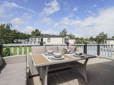 A patio deck with a wicker dining table set with plates bowls and flowers overlooking mobile homes at Yealands 27 South Lakeland Leisure Village near Carnforth