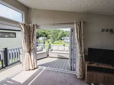 A living room with curtains open to a deck with outdoor seating at Yealands 27 South Lakeland Leisure Village near Carnforth
