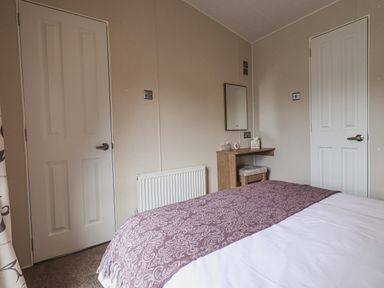 A bedroom with a bed covered by a patterned blanket two white doors a wooden dressing table with a mirror and a cushioned stool at Yealands 27 South Lakeland Leisure Village near Carnforth