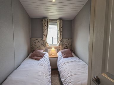 A small bedroom with two single beds separated by a nightstand with a lamp and a window with curtains at Yealands 27 South Lakeland Leisure Village near Carnforth