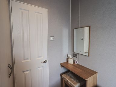 A small dressing table with a mirror tissue box and a cushioned stool next to a closed white door at Yealands 27 South Lakeland Leisure Village near Carnforth
