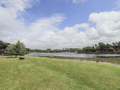 A grassy area next to a lake with wooden cabins and trees under a partly cloudy sky at Yealands 27 South Lakeland Leisure Village near Carnforth