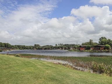 A lake with grass and reeds in the foreground and wooden cabins along the shore at Yealands 27 South Lakeland Leisure Village near Carnforth