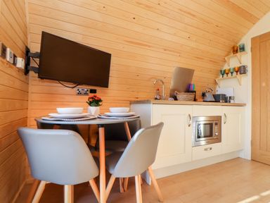 A small kitchen and dining area with a wall-mounted TV a round table set with bowls and plates and a kitchenette with a microwave and sink at Sea View in St Buryan