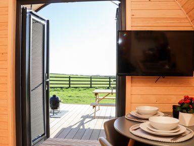 An open door leading to a deck with a picnic table and a green field beyond a black fence with a table set with bowls plates and placemats inside the wooden room at Sea View in St Buryan