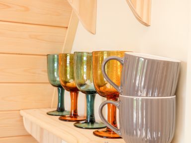 Two gray ceramic mugs stacked beside four colorful glass goblets on a wooden shelf at Sea View in St Buryan