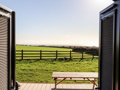 View of green fields with a wooden fence and a picnic table outside through open doors at Sea View in St Buryan