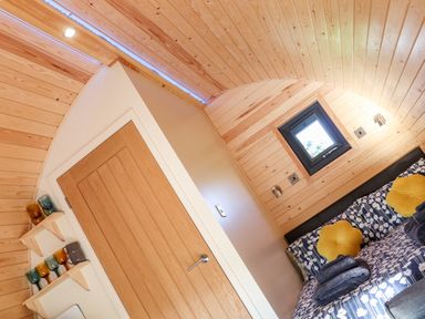 A small bedroom with wooden walls and ceiling a bed with patterned bedding and yellow pillows and a wooden door with shelves holding mugs at Sea View in St Buryan