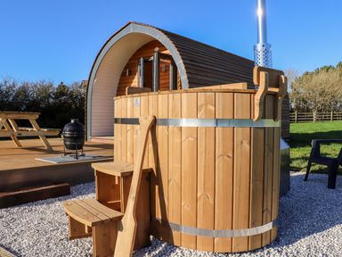 An outdoor wooden hot tub with steps and a wooden paddle next to a curved wooden cabin with open doors at Sea View in St Buryan