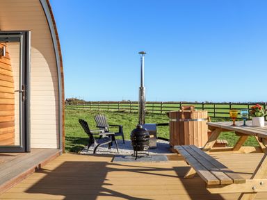 A wooden deck with a picnic table and chairs a hot tub and a barbecue grill in a fenced grassy field at Sea View in St Buryan