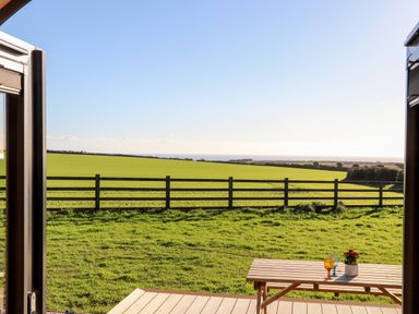 A view of a green field with a wooden fence and a wooden picnic table with glasses and a flowerpot on a deck at Sea View in St Buryan