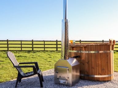 A wooden hot tub with metal stove and two glasses beside a black plastic chair outside in a fenced grassy area at Sea View in St Buryan