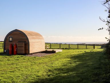 A wooden pod on a grass field with a wooden fence and sea in the background at Sea View in St Buryan