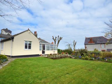 A single story house with a garden and flower beds under a cloudy sky at Hafod Cottage in Cemaes Bay