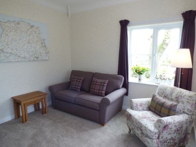 A living room with a purple sofa and plaid cushions a floral armchair a lamp and a window with purple curtains at Hafod Cottage in Cemaes Bay
