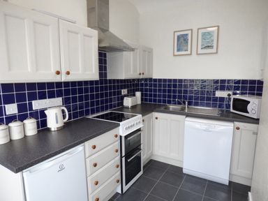 A kitchen with white cabinets blue tiled backsplash a stove dishwasher microwave kettle and canisters at Hafod Cottage in Cemaes Bay