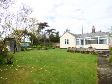 A garden with grass trees a small stone building a car and a cream house with a tiled roof at Hafod Cottage in Cemaes Bay