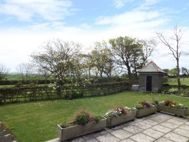 A garden with flower boxes on a paved area a small stone building and trees in the background at Hafod Cottage in Cemaes Bay