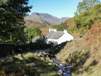 A white house near a small stream with trees and mountains in the background at Brookside in Grizebeck