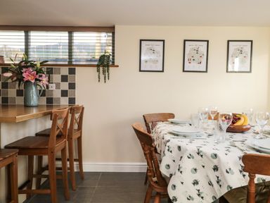 A dining area with wooden chairs around a table with a patterned tablecloth and a kitchen counter with wooden bar stools and a vase with flowers at Brookside in Grizebeck