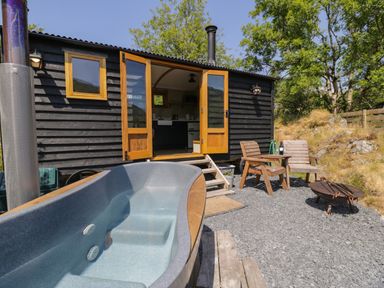 An outdoor area with a wooden hut opening to a kitchen an empty hot tub wooden chairs with a table holding a wine bottle and glass and a fire pit at Llethr Shepherds Hut in Llanbedr
