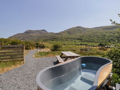 An outdoor area with a hot tub and wooden picnic table with mountains in the background at Llethr - Shepherds Hut in Llanbedr