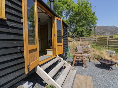 An outdoor seating area with wooden chairs and fire pit next to a black wooden building with open doors at Llethr - Shepherds Hut in Llanbedr