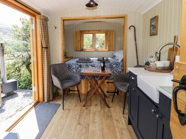 An open tiny house with a wooden floor a small wooden table with two chairs a bed with window shutters and a kitchenette with a sink and countertop at Llethr - Shepherds Hut in Llanbedr