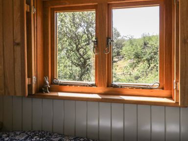 A wooden window with metal latches and a small rabbit figurine on the sill overlooking trees at Llethr - Shepherds Hut in Llanbedr