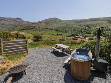 An outdoor area with a wooden hot tub a picnic bench a fire pit and mountains in the background at Llethr - Shepherds Hut in Llanbedr