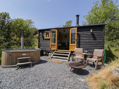 A small wooden cabin with open doors a hot tub wooden chairs a fire pit and trees at Llethr - Shepherds Hut in Llanbedr