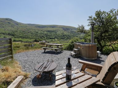 An outdoor seating area with a wooden bench a bottle of wine and two glasses a fire pit a wooden picnic table and a wooden hot tub with hills in the background at Llethr - Shepherds Hut Llanbedr