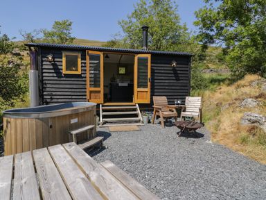 An outdoor area with a black wooden cabin having open double doors a wooden hot tub wooden chairs table and fire pit at Llethr - Shepherds Hut in Llanbedr