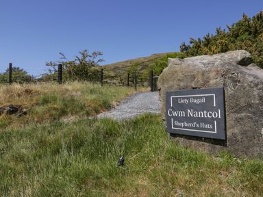 A gravel path lined with a fence and grass next to a large rock with a sign for Cwm Nantcol Shepherds Huts