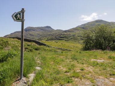 A grassy walking trail with a pedestrian signpost and mountains in the background at Llethr - Shepherds Hut in Llanbedr